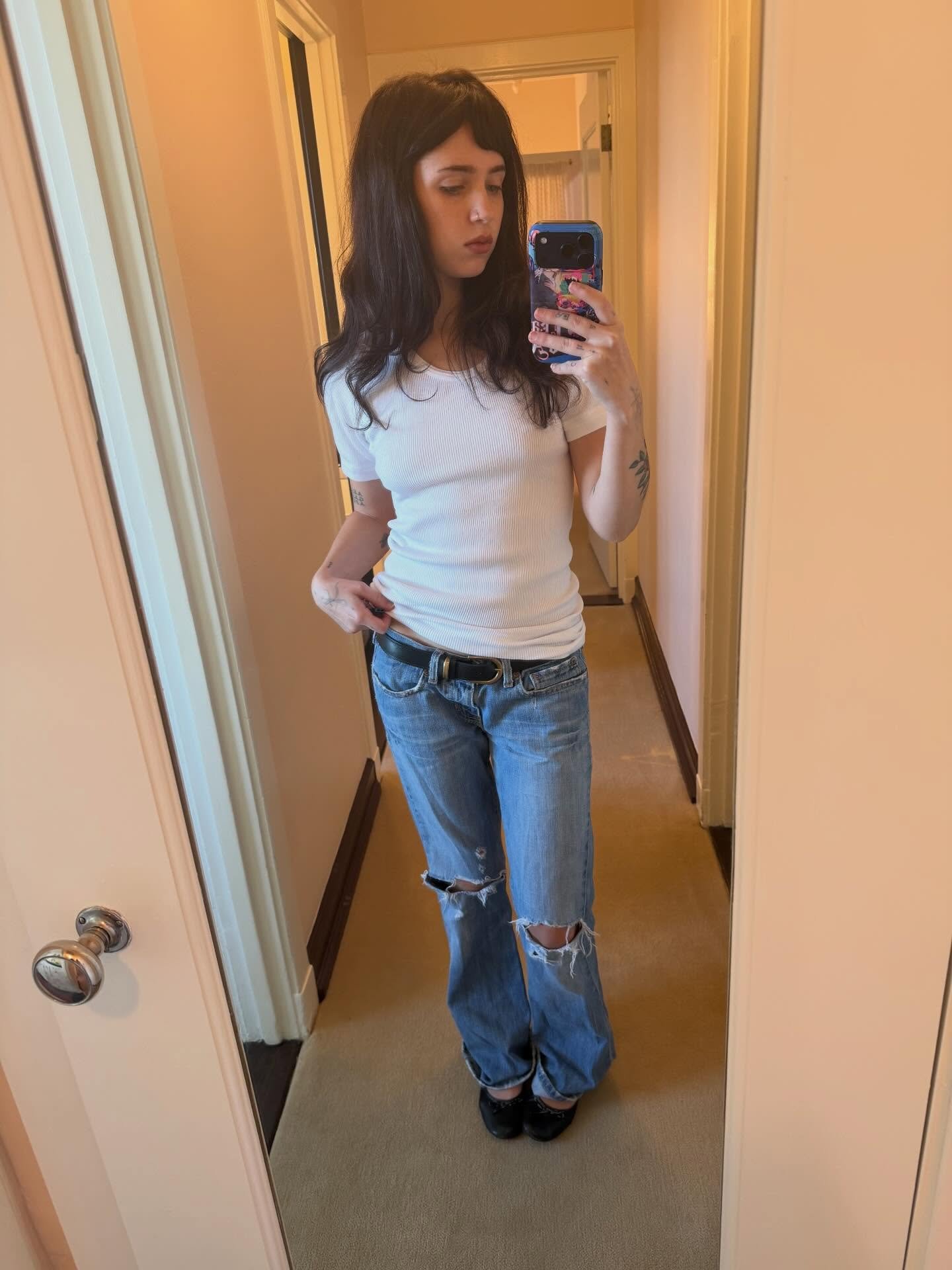 Clairo is wearing ripped skinny jeans, black belt, white T-shirt, and black ballet flats