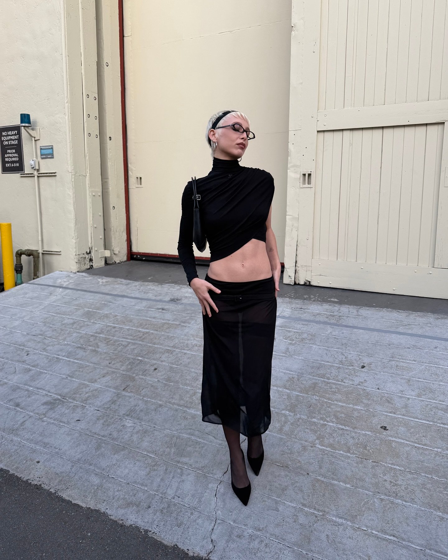 Emma Chamberlain is wearing a sheer black skirt, tights, a black long sleeve, and pointed-toe heels